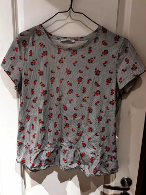 Blommig randig blus med volang - Söt blus med smala blå och vita ränder och små röda blommor över hela tyget. Kortärmad modell med rund halsringning och volangkant nedtill.