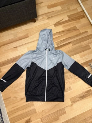 Nike vindjacka med huva, svart/grå - Snygg vindjacka från Nike i svart och ljusgrått med dragkedja framtill och huva med dragsko. Jackan har två fickor och den klassiska Nike-loggan på bröstet. Perfekt för blåsiga dagar och sportiga outfits.