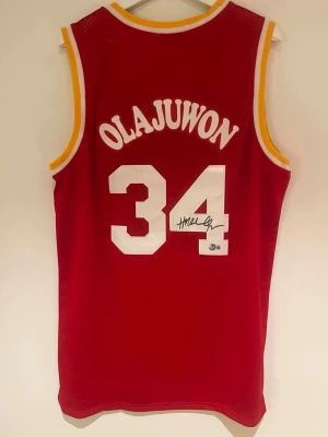 Signerad tröja utav Hakeem Olajuwon - Beckett Certifierad - Nu har du chansen att lägga vantarna på en riktigt ikonisk samlarpryl! Denna röda baskettröja med nummer 34 är personligen signerad av NBA-legenden Hakeem Olajuwon – en av de bästa centrarna genom tiderna.  Autografen är Beckett-certifierad Tydlig och snygg signatur på siffran Perfekt för samlare eller som exklusiv present Klassisk design – riktigt snygg att rama in och hänga upp  Hakeem “The Dream” Olajuwon är Hall of Famer, 2× NBA-mästare och en av sportens mest respekterade spelare!