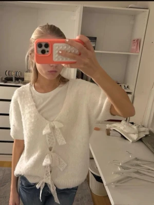 Vit kofta med rosettknäppning - Söt vit kofta med korta ärmar och tre dekorativa rosetter framtill som knäppning. Koftan har en fluffig, stickad struktur och djup v-ringning. Perfekt att styla över en t-shirt och jeans för en trendig look.