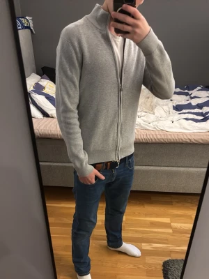 Ljusgrå ziptröja från Bläck - Säljer en stilren ljusgrå ziptröja med långa ärmar. Tröjan har ribbade muddar vid ärmslut och nederkant samt dragkedja hela vägen. Perfekt att slänga på sig över en t-shirt för en clean look.