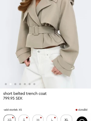 Kort beige trenchcoat med bälte - Trendig kort trenchcoat i beige med breda slag, markerad midja och matchande bälte. Jackan har stora manschetter med knappdetalj och en klassisk krage. Perfekt för dig som vill ha en modern twist på trenchcoaten.