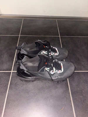 Nike reacts vision svarta sneakers - Nike reacts vision sneakers i svart och grått med transparent sula och svarta snören. Ovandelen är i meshmaterial med vita och röda detaljer vid snörningen. Snygg och sportig design med dragflik bak för enkel påtagning.