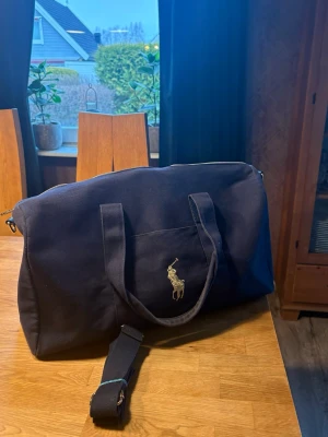 Svart dufflebag från Polo Ralph Lauren - Snygg svart dufflebag från Polo Ralph Lauren i slitstark canvas med klassisk guldfärgad logga framtill. Väskan har både handtag och justerbar axelrem samt dragkedja i metall. Perfekt storlek för kortare resor och har en enkel, stilren design. Använd ett fåtal gånger och är i bra skick pris ej hugget i sten