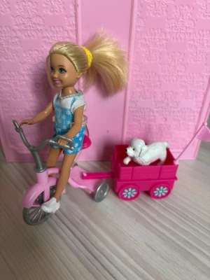 Docka med cykel och hundvagn - Säljer en blond docka med blåa hängselshorts och vit topp. Dockan cyklar på en rosa trehjuling med gråa detaljer och har en rosa vagn kopplad bakom sig. I vagnen sitter en liten vit hundfigur. Perfekt för dig som gillar roliga docktillbehör och lekset.