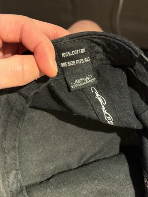 Svart Ed Hardy keps med strasstenar - Säljer en svart keps från Ed Hardy by Christian Audigier. Kepsen är dekorerad med färgglada strasstenar, döskalle och texten 'Love Kills Slowly' framtill. Broderad logga på skärmen och justerbar passform. Tillverkad i 100% bomull.