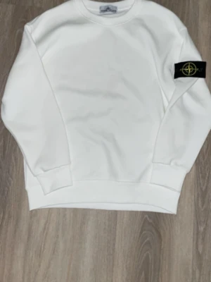 Vit sweatshirt från Stone Island - Säljer en clean vit sweatshirt från Stone Island med den klassiska svarta logotypen på vänster ärm. Tröjan har rund hals, ribbade muddar och är tillverkad i mjukt bomullsmaterial. Perfekt för en stilren och avslappnad look.