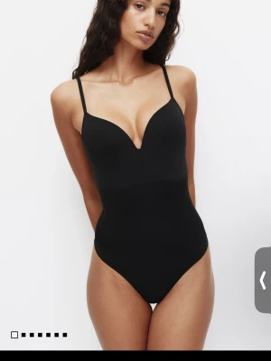 Svart shaping-body med tunna band - Snygg svart shaping-body med tunna, justerbara axelband och djup urringning. Tillverkad i ett slätt, stretchigt material som formar kroppen och ger en smickrande siluett. Perfekt under tighta plagg för en sömlös look.