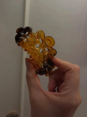 Selór likande hårklämma  - Säljer en stor hårklämma i plast med blommönster. Klämman är bärnstensfärgad med mörkare bruna detaljer i kanterna och har ett genomskinligt, glansigt utseende. Perfekt för att snabbt sätta upp håret med en snygg touch.
