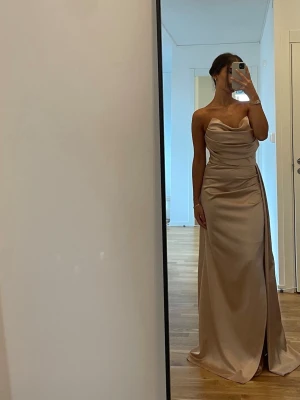 Champagnefärgad långklänning omlott - Elegant långklänning i champagnefärg med omlottdetalj och slits framtill. Klänningen är axelbandslös och har en draperad överdel som ger en lyxig känsla. Perfekt för speciella tillfällen där du vill sticka ut. Köpt för 2500 kr. Använd 1 gång. 