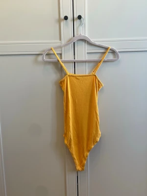 Gul ribbad body från Bik Bok - Säljer en gul body från Bik Bok i ribbat stretchigt material. Modellen har smala axelband och en enkel, stilren design som sitter tajt mot kroppen. Perfekt att styla med jeans eller kjol för en trendig look.