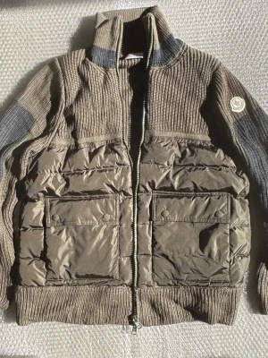 Olivgrön cardiganjacka Moncler - Snygg olivgrön cardiganjacka från Moncler med stickade partier och vadderade paneler. Jackan har två stora fickor framtill med knappar, hög krage och dragkedja. Materialmix av stickat tyg och glansigt nylon ger en cool kontrast. Moncler-logga på ärmen.