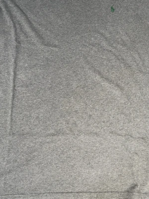 Grå V-ringad tröja Polo Ralph Lauren - Snygg gråmelerad V-ringad tröja från Polo Ralph Lauren. Klassisk passform med diskret grön logga broderad på bröstet. Tillverkad i mjukt material som känns skönt mot huden. Perfekt till jeans eller över en skjorta.