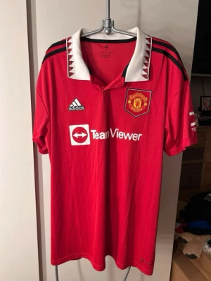 Manchester United tröja Adidas röd - Röd Manchester United matchtröja hemma, säsongen 22/23, från Adidas med vit krage och svarta detaljer. Broderad klubbmärke på bröstet, TeamViewer sponsortryck och klassiska Adidas-ränder på axlarna. Tillverkad i lätt och ventilerande polyester, perfekt för fotbollsträning eller match.   Bara använd en gång, så den är fortfarande så gott som ny! Ligger även ute på Vinted.