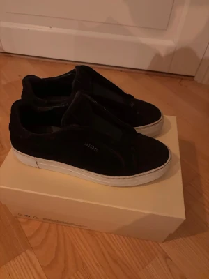 Svarta sneakers från Axel Arigato - Snygga svarta sneakers från Axel Arigato med minimalistisk design och vit sula. Skorna är låga och har en stilren look utan synliga snören, tillverkade i mocka för en lyxig känsla. Perfekta för dig som gillar clean streetstyle.