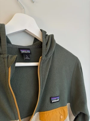 Fleecehoodie från Patagonia - Säljer en grön och beige fleecehoodie från Patagonia med dragkedja och huva.  Använd fåtal gånger och har ingen defekter. Passar mellan 155/165 cm. Perfekt nu inför våren! 