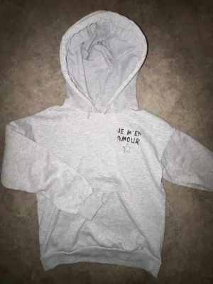Grå hoodie med tryck 'Je m'en amour' - Snygg ljusgrå hoodie med svart texttryck 'Je m'en amour' och en stjärna både fram och bak. Hoodien har stor huva och ribbade muddar vid ärmar och nederkant. Perfekt för en chill och avslappnad stil. Materialet känns mjukt och bekvämt, troligen bomullsblandning.
