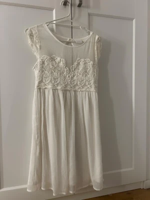 Vit spetsklänning med snörning - Romantisk vit klänning med spetsdetaljer över bysten och axlarna. Klänningen har en luftig kjol i lätt tyg, rund halsringning och snörning i ryggen. Perfekt för sommarens alla tillfällen och ger en bohemisk känsla.