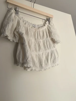 Vit offshoulder topp från Zara - Söt vit offshoulder topp från Zara i storlek 164. Toppen har puffiga korta ärmar, volangdetaljer och en luftig, krinklad struktur. Perfekt för en trendig och avslappnad look med romantiska vibes.