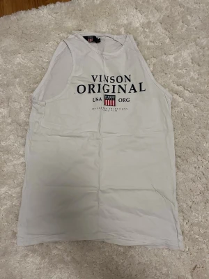 Vit linnetopp från Vinson Original - Säljer en vit linnetopp från Vinson Original med tryck i svart och rött framtill. Toppen är ärmlös och har rund halsringning. Perfekt för en avslappnad och sportig stil.