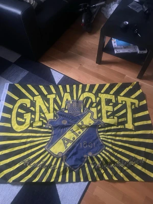 AIK supporterflagga GNAGET - Stor supporterflagga i gult och svart med texten GNAGET och AIK:s klubbmärke i mitten. Flaggan har strålande linjer från mitten och texten 'Allmänna Idrottsklubben' längst ner. Perfekt för AIK-fans att visa sitt stöd.