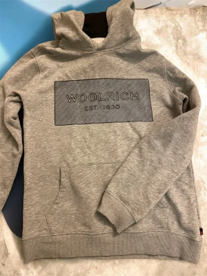 Grå hoodie från Woolrich - Snygg grå hoodie från Woolrich med stor logga och texten 'EST. 1830' på bröstet. Klassisk känguruficka framtill och ribbade muddar vid ärmslut och nederkant. Tillverkad i mjukt material och har en skön huva. Perfekt för en avslappnad stil.