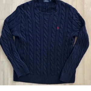 Mörkblå stickad tröja Polo Ralph Lauren - Snygg mörkblå stickad tröja från Polo Ralph Lauren med klassiskt kabelstickat mönster och rund hals. Tröjan har långärmade ribbade muddar och en liten röd broderad logga på bröstet. Perfekt för dig som gillar stilren och tidlös design.