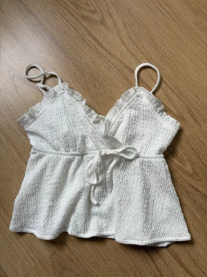 Vit volang topp - Söt vit linnetopp med omlottdesign, tunna axelband och volangdetaljer vid ringningen. Toppen har ett knyt i midjan och är tillverkad i ett lätt, strukturerat material. Perfekt för varma dagar och enkel att styla 