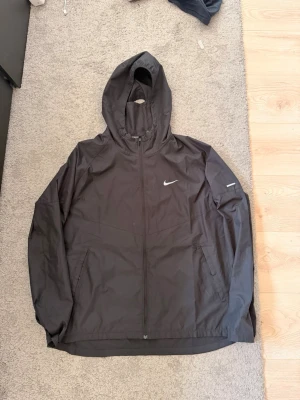 Svart Nike vindjacka med huva - Snygg svart vindjacka från Nike med huva och dragkedja framtill. Jackan har två fickor med dragkedja och en vit Nike-logga på bröstet samt en reflexdetalj på ärmen. Tillverkad i lätt och vindtätt material, perfekt för blåsiga dagar.