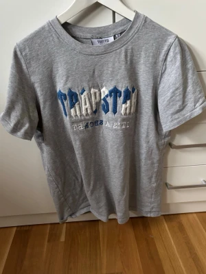 Trapstar t shirt  - Säljer en grå t-shirt från Trapstar med broderad logga i blått och vitt framtill. T-shirten har rund halsringning och korta ärmar. Tillverkad i mjuk bomull för skön passform och streetwear-vibe.