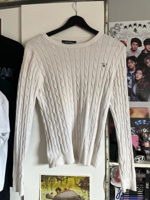 Vit kabelstickad tröja från Gant - Kabelstickad vit tröja från Gant med rund halsringning och diskret logga broderad på bröstet. Tröjan har långa ärmar och ribbade muddar vid ärmslut och nederkant. Klassisk och stilren design som passar till mycket.