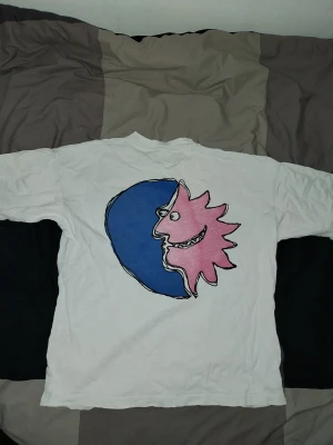 District 46 T shirt  - Vit t-shirt från District 46 med ett stort grafiskt tryck av en blå måne och en rosa sol på ryggen. Framtill finns en liten logga på bröstet. T-shirten har rund hals och korta ärmar. Perfekt för dig som gillar unika prints och streetwear vibes.