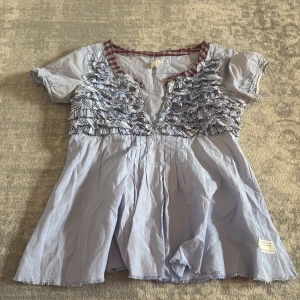 Ljusblå blus med volangdetaljer - Supersöt ljusblå blus från Odd Molly med korta ärmar och volangdetaljer över bröstet. Blusen har rutigt kantband vid halsringningen och en lätt v-ringning framtill. Perfekt för en trendig och avslappnad look.