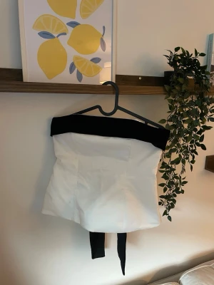 axelbandslös topp med rosett från ZARA - Snygg axelbandslös topp i vitt med bred svart kant upptill och stor svart rosett bak. Toppen har bandeau-modell och långa band som hänger ner från rosetten. Perfekt för dig som vill sticka ut med en elegant och trendig look.
