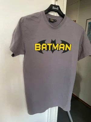 Batman t-shirt i grå bomull - Snygg grå t-shirt med klassiskt Batman-tryck i svart och gult på bröstet. Tillverkad i mjuk bomull och har en normal passform med korta ärmar. Perfekt för dig som gillar superhjältar och vill ha en cool vardagslook.