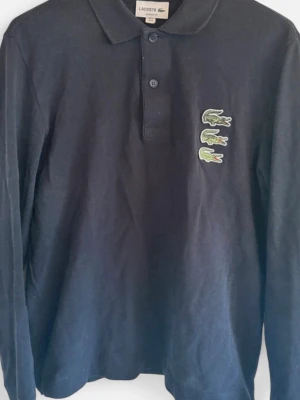 Marinblå långärmad piké från Lacoste - Klassisk marinblå långärmad piké från Lacoste med tre gröna krokodillogos på bröstet. Pikén har krage och knappar framtill. Perfekt för dig som gillar stilrena plagg med en twist. Perfekt till våren/sommaren