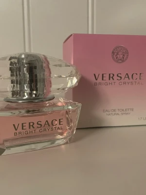 Versace Bright Crystal Eau de Toilette - Versace Bright Crystal Eau de Toilette är en lyxig parfym med fräsch och blommig doft. Flaskan är elegant i genomskinligt glas med rosa inslag och har ett kristalliknande lock. Förpackningen är rosa och vit med Versace-loggan och stilren text.