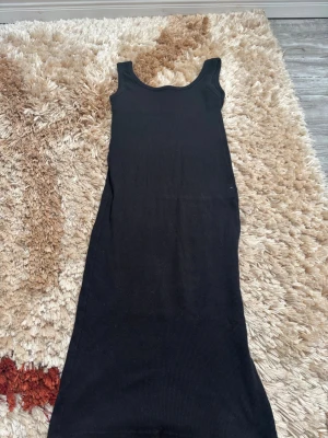 Svart ribbad långklänning - En svart, ribbad långklänning med breda axelband och rundad halsringning. Klänningen har en enkel och stilren design som passar perfekt till många olika tillfällen. Materialet är stretchigt och följsamt för en bekväm passform.