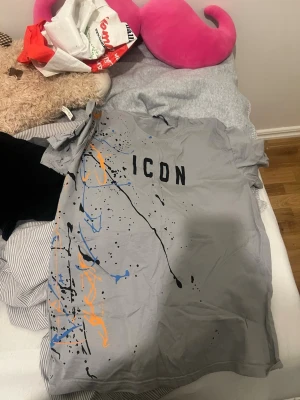 Grå ICON t-shirt från Dsquared2 - Säljer en grå t-shirt från Dsquared2 med trycket 'ICON' på bröstet och färgstänk i blått, orange och svart över framsidan. T-shirten är kortärmad och har en normal passform. Perfekt för dig som vill sticka ut med en unik och trendig look.