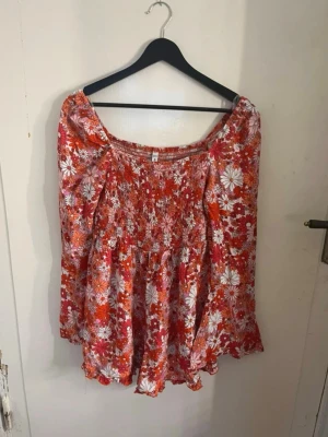 Röd blommig kort shortsdress med volang - Supersöt kort shortsklänning med röd och vit blommig print, smockad överdel och volangkant. Den har långa ärmar och en offshoulder-modell som ger en avslappnad och trendig look. Perfekt för sommarens alla tillfällen! Det är alltså en kortklänning men kjolen är shorts. Den är aldrig använd, isåfall bara nån enstaka gång. Kan bli snyggt att styla med ett par boots med mera. 