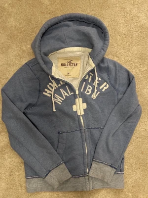 Blå hoodie från Hollister Malibu (Reserverad) - Säljer en blå hoodie från Hollister Malibu med dragkedja och vita detaljer. Hoodien har en stor huva med dragsko, två fickor framtill och ribbade muddar vid ärmar och nederkant. Tryckt text på bröstet och mjukt material på insidan.
