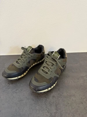 Valentino camo sneakers - Snygga sneakers från Valentino i militärgrönt camomönster med svarta och olivgröna detaljer. Skorna har lågt skaft, snörning och coola nitar längs sulan. Materialmix av textil och läder ger en exklusiv känsla och streetwear-vibe.