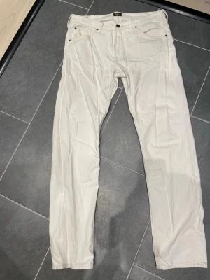Vita raka jeans från Lee - Säljer ett par vita jeans från Lee med rak passform och klassisk femficksdesign. Jeansen har metallknapp och nitar, samt Lee-logga på linningen. Tillverkade i kraftigt bomullstyg som ger en clean och stilren look.