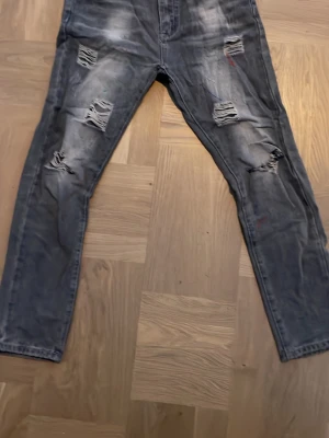 Grå slitna jeans med hål - Säljer ett par grå jeans med slitningar och hål på framsidan. Jeansen har en slim fit och detaljer med färgstänk för en cool, avslappnad look. Bakfickor och klassisk femficksmodell. Perfekta för dig som gillar streetstyle.