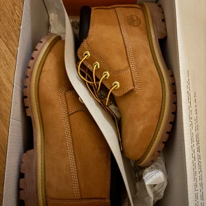 Timberland klassiska kängor i nubuck - Säljer ett par klassiska Timberland kängor i ljusbrun nubuck med robust gummisula och guldfärgade metalldetaljer. Kängorna har vita kontrastsömmar, högt skaft och logga på sidan. Perfekta för dig som vill ha en ikonisk streetwear-look. EU 37.5