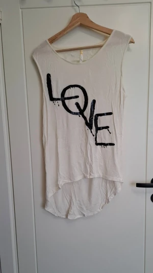 Vit ärmlös topp från Ocasion - Säljer en vit ärmlös topp från Ocasion med svart texttryck 'LOVE' framtill. Toppen har rund halsringning och längre bakstycke för en cool, avslappnad look. Perfekt till jeans eller shorts för en chill stil.