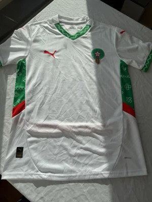 Marocko fotbollströja Puma vit/grön - Säljer en officiell Marocko fotbollströja från Puma i vitt med gröna och röda detaljer. Tröjan har korta ärmar, grönt mönster på sidorna och röd Puma-logga på bröstet. Tillverkad i lätt och ventilerande material med DryCell-teknologi.
