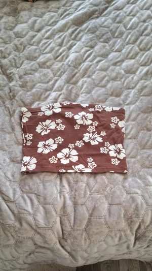 Brun blommig tubtopp - Säljer en brun tubtopp med vita hibiskusblommor. Toppen är axelbandslös och har en rak, enkel passform. Perfekt för sommaren och ger en chill strandvibe. Materialet känns mjukt och stretchigt, vilket gör den bekväm att bära hela dagen.