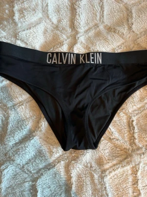 Svart bikiniunderdel Calvin Klein M - Svart bikiniunderdel från Calvin Klein med bred resår i midjan och tydlig logga framtill. Snygg och enkel design som passar perfekt till stranden eller poolen. Tillverkad i mjukt och stretchigt material för skön passform.