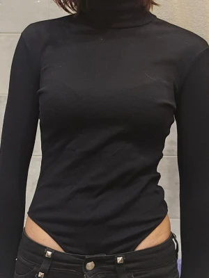 Svart polotopp med lång ärm - ❗️Skriv innan köp❗️Svart, tight polotopp med lång ärm och ribbad struktur. Toppen har hög hals och är figurnära. Materialet känns stretchigt och bekvämt. Ärmarna har tumhål men finns dock flera hål efter användning. Annars är bodyn i ganska bra skick. Det finns knappar därnere och kan täcka hela överkroppen om man skulle vilja det. 